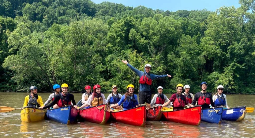 a group of people wearing safety gear sit in several canoes and smile for the photo.